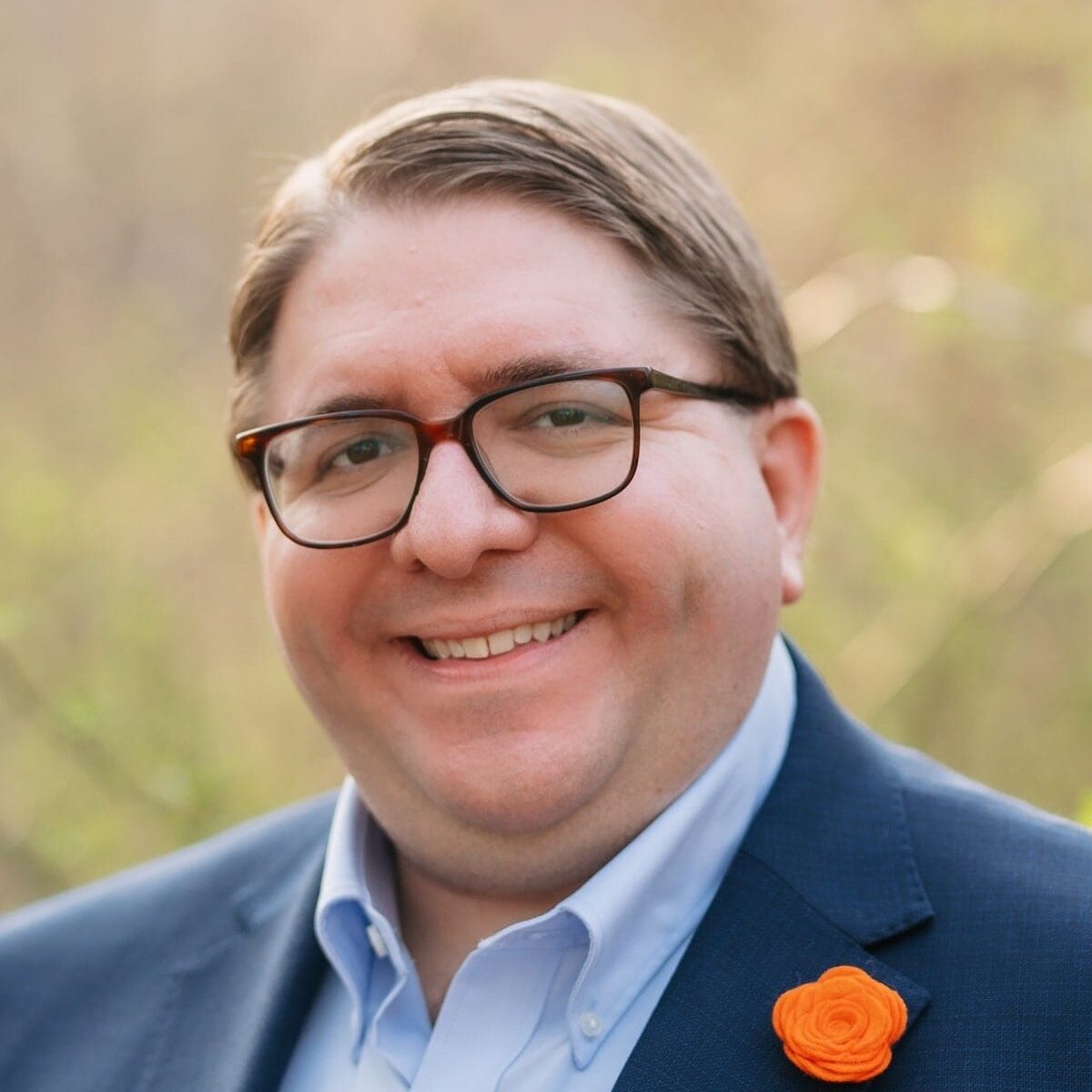 Smiling man wearing glasses, a navy suit, light blue shirt, and orange boutonniere outdoors